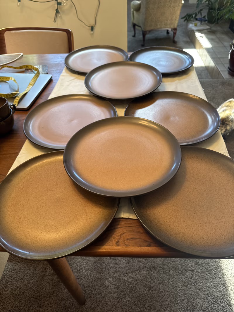 Eight brown ceramic plates are arranged in a circular pattern on a wooden dining table with a table runner, in a sunlit room.
