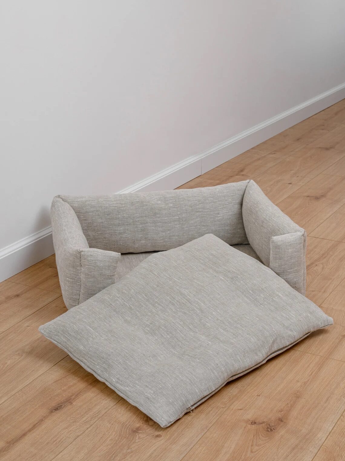 A light gray pet bed with a detached cushion sits on a light wood floor against a plain white wall.