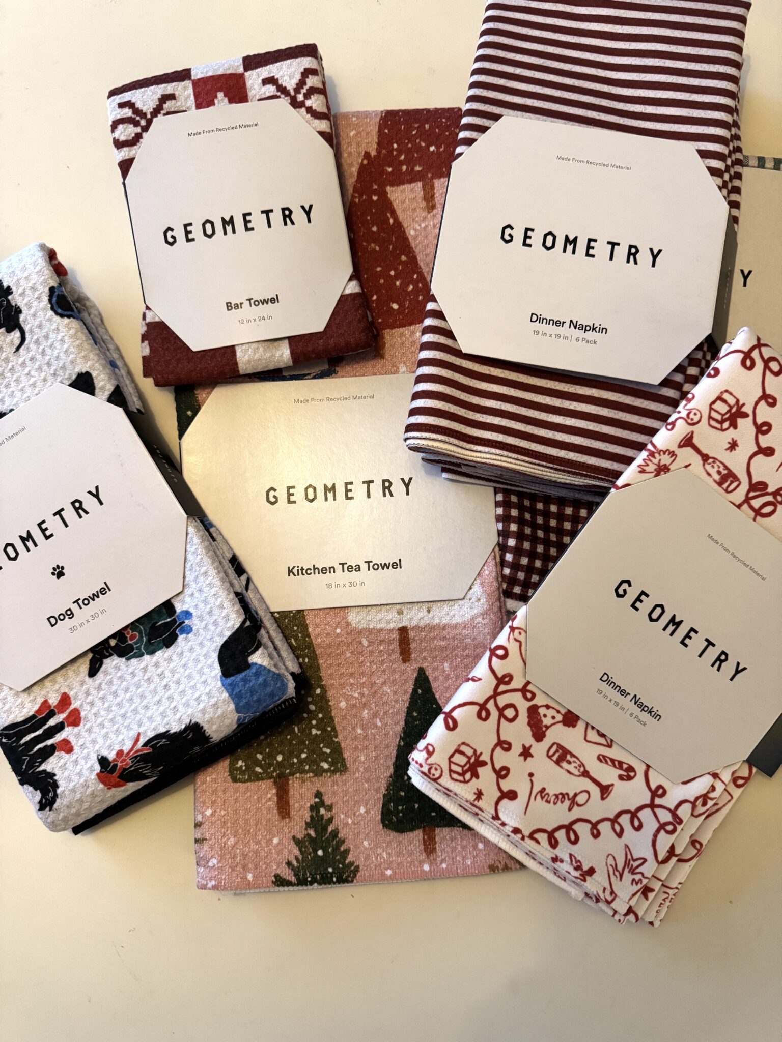 Several Geometry brand textiles, including bar towels, kitchen tea towels, dog towels, and dinner napkins, are displayed on a flat surface in various patterns and colors.