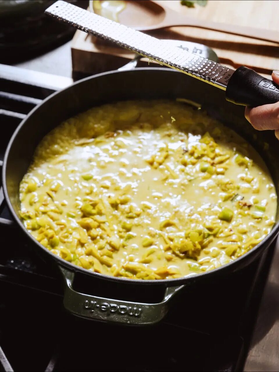 A hand grates cheese or zest over a pan filled with a creamy mixture of corn and sauce, cooking on a stovetop.