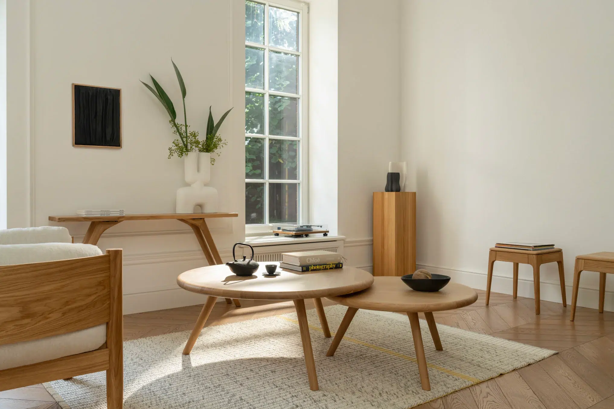 A minimalist living room with light wood furniture, a large window, circular coffee tables, books, decorative vases, and neutral tones.