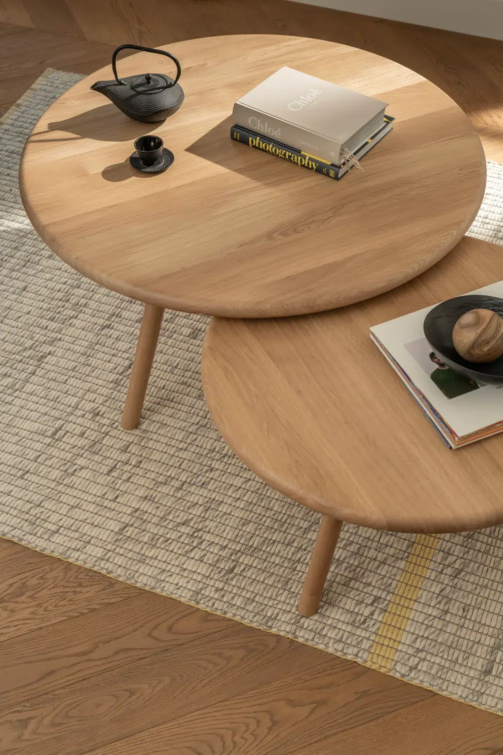 Two round wooden coffee tables on a textured rug, each holding books and decorative objects, with sunlight casting soft shadows across the scene.