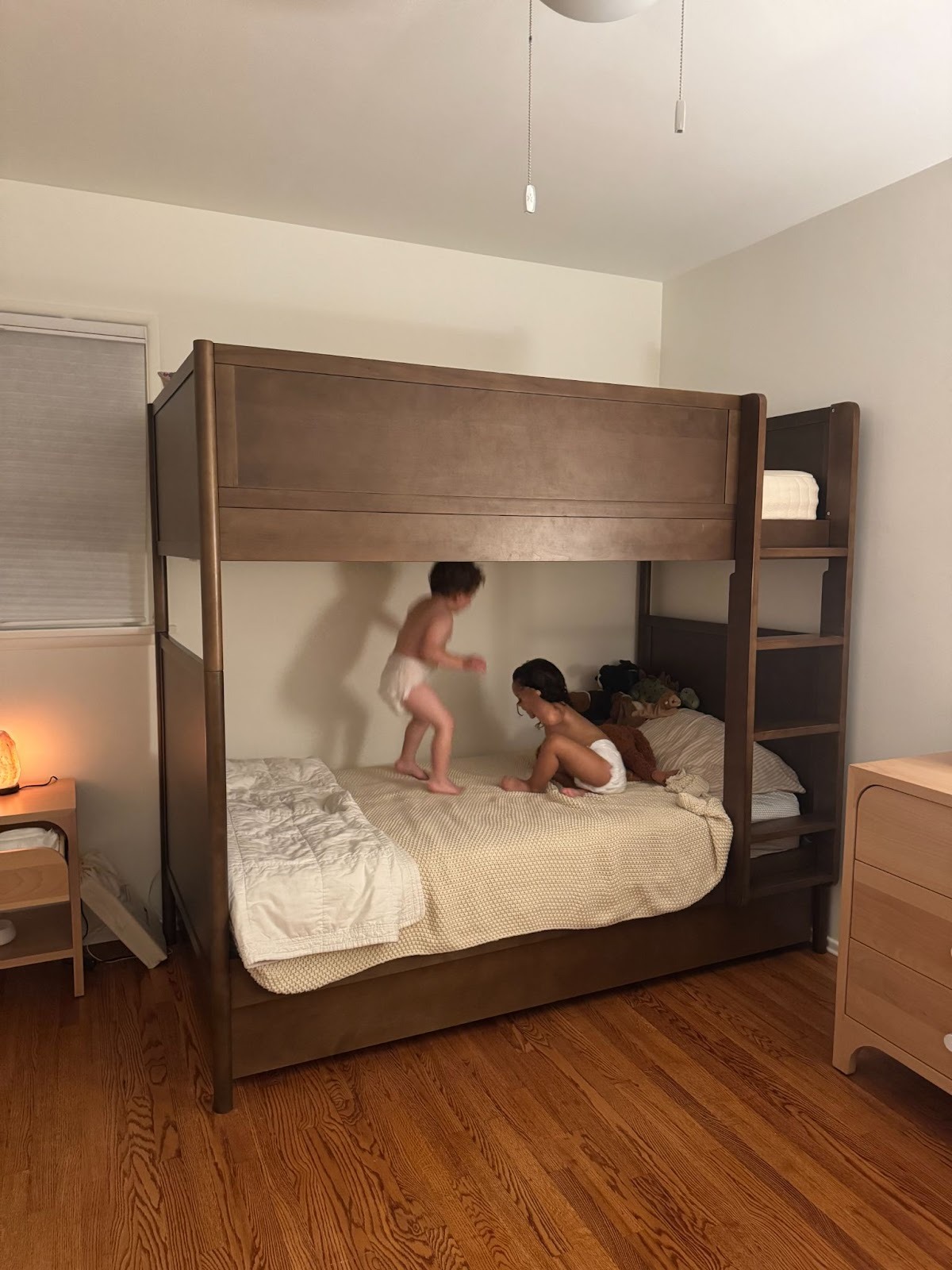 Two young children in diapers are on a wooden bunk bed in a bedroom with hardwood floors and minimal furnishings. One child is jumping on the lower bunk while the other lies on the bed.