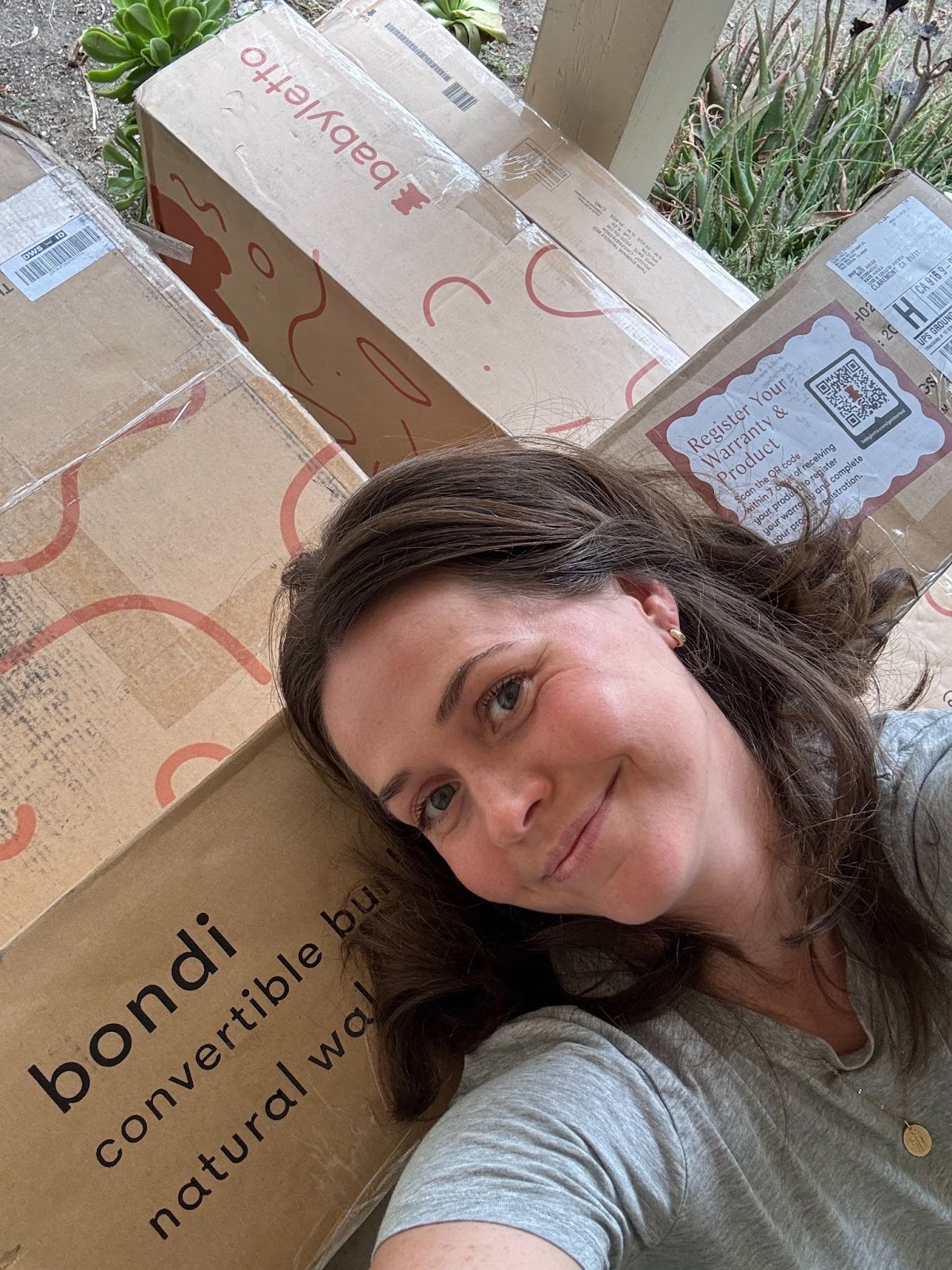 A woman lies next to several large cardboard boxes, including ones labeled "bondi convertible bunk" and "babyletto.