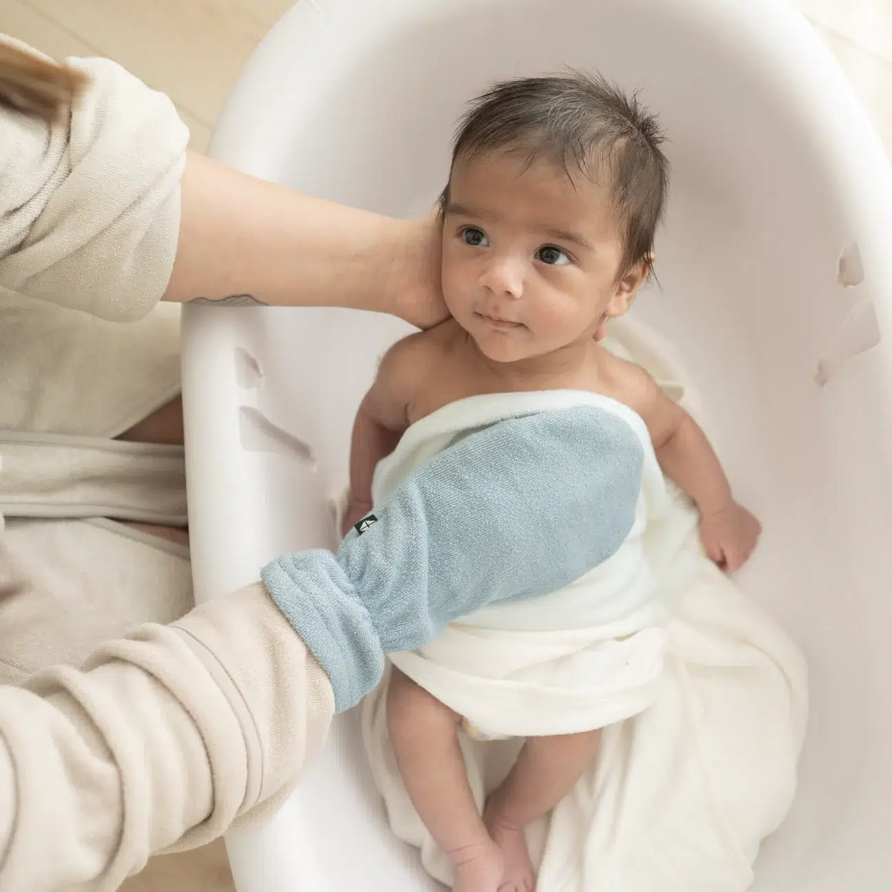 An adult supports and washes a baby wrapped in a white towel inside a white baby bathtub, using a light blue wash mitt.