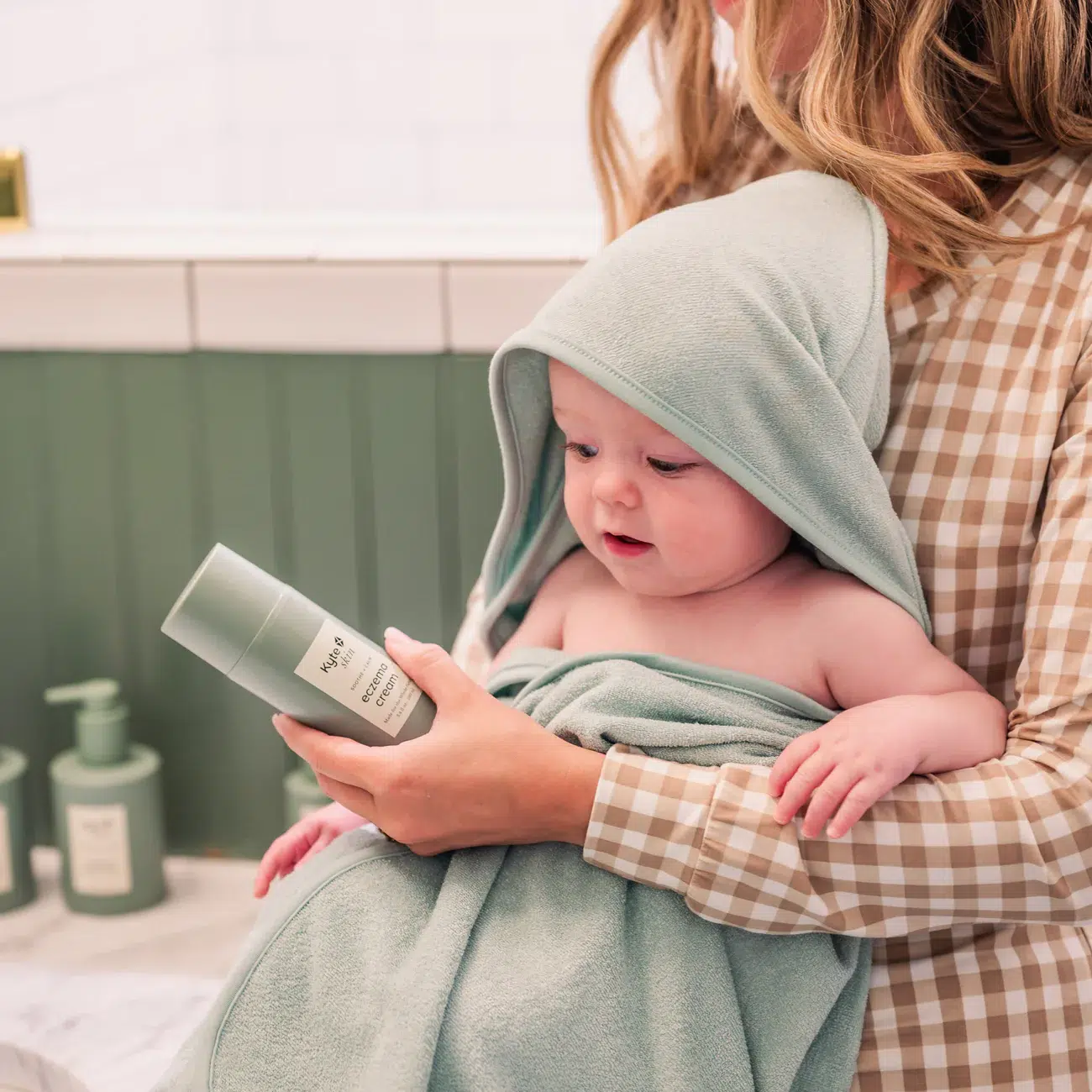 An adult holding a baby wrapped in a green hooded towel shows the baby a skincare product in a bathroom.