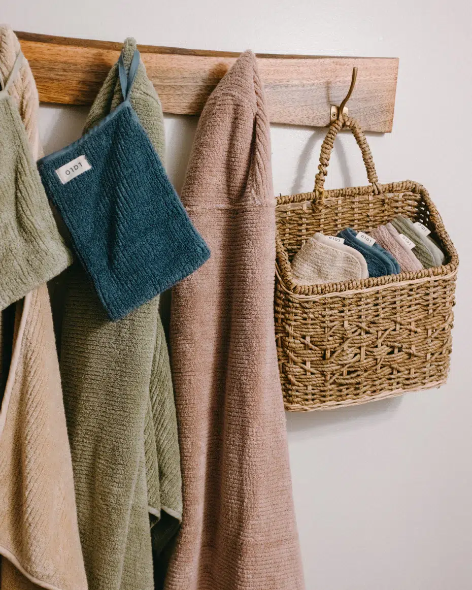 A wooden rack holds towels in muted colors, a blue washcloth, and a woven basket filled with folded washcloths against a white wall.
