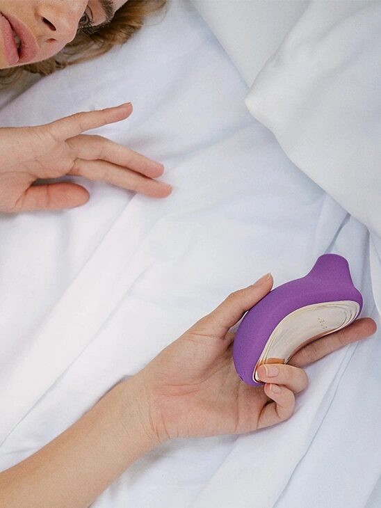 A person lying on a bed holds a small, purple, curved object in their hand near white sheets.