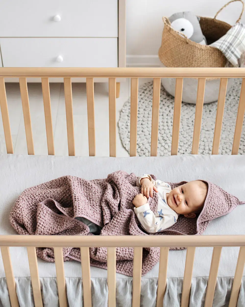 A baby lies in a wooden crib, wrapped in a mauve knitted blanket, with a dresser and a basket holding a stuffed animal in the background.