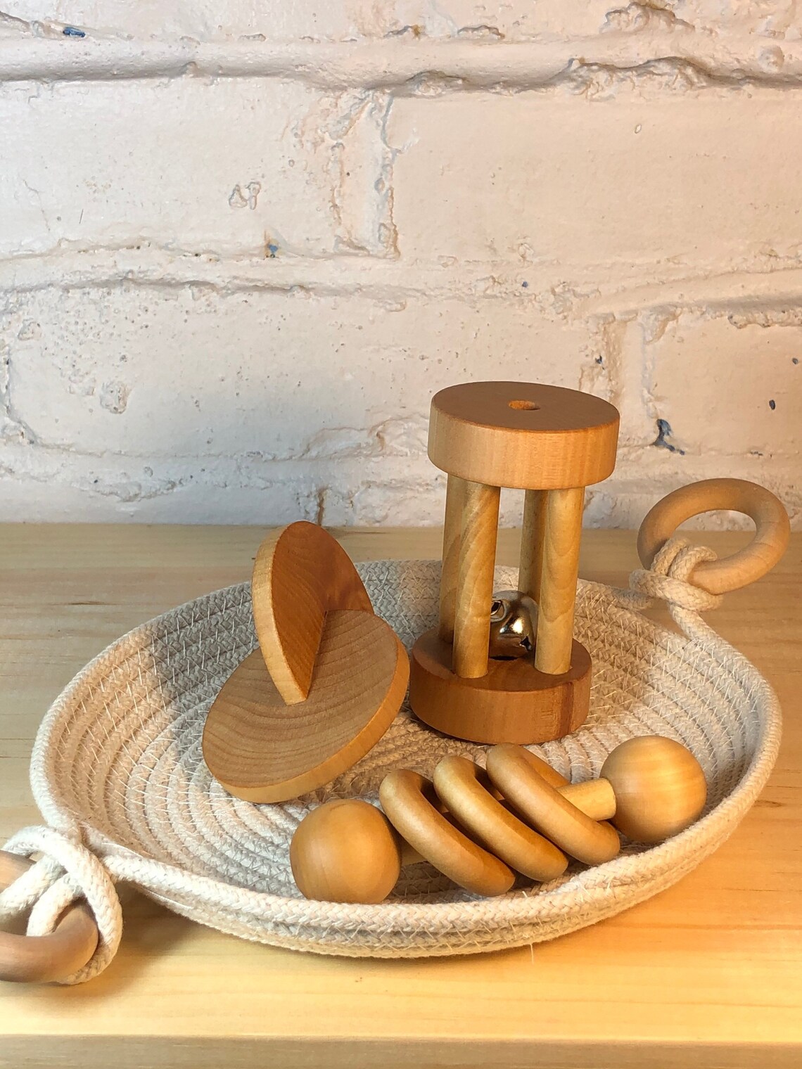 A woven basket on a wooden surface holds three wooden baby toys: a disc set, a rattle with rods and a bell, and a ring stacker. A white brick wall is in the background.