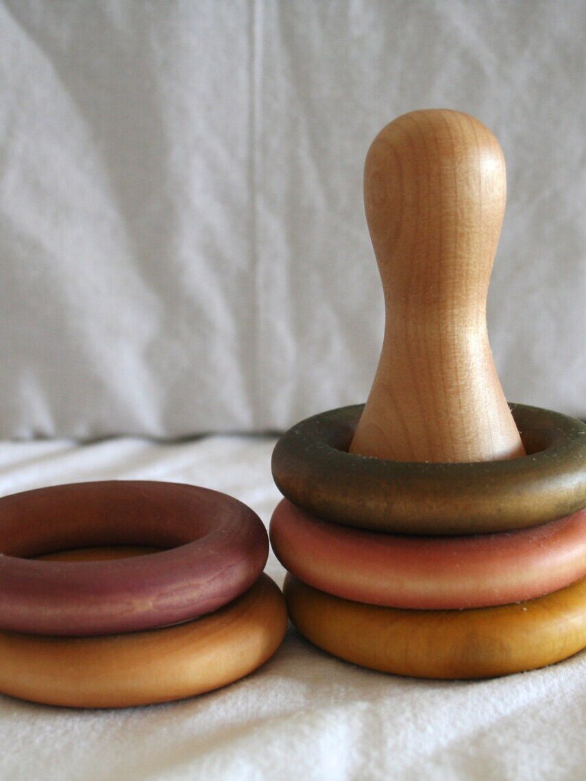 A wooden stacking toy with a base post and four colored rings, three stacked on the post and one beside it, placed on a white fabric surface.