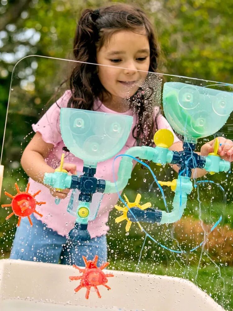 A young girl plays outside with a water activity wall featuring funnels, gears, and spinning wheels on a clear panel.