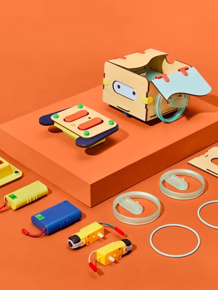 Various colorful electronic and mechanical components, including plastic parts, wires, and a wooden box, are arranged neatly on an orange background.