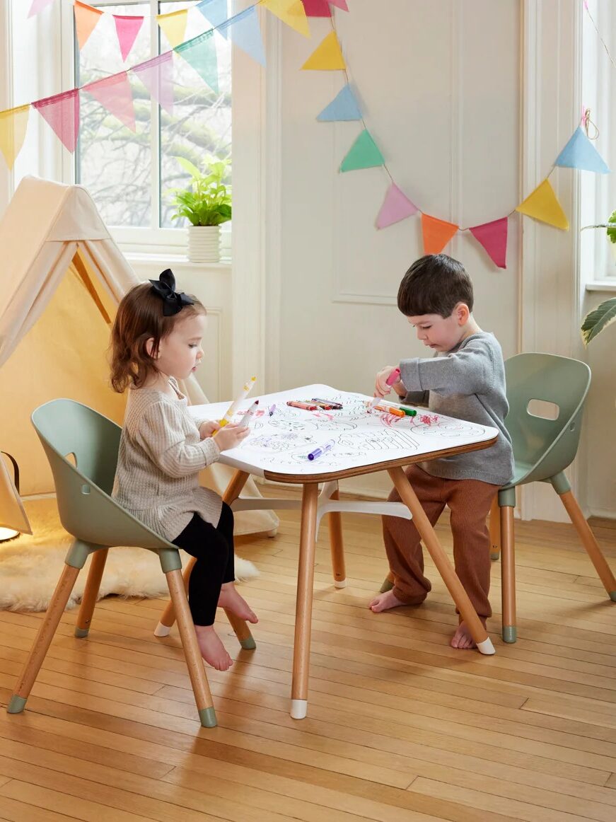 Two young children sit at a small table coloring with markers in a playroom decorated with a tent, colorful pennant banner, and wooden floor.