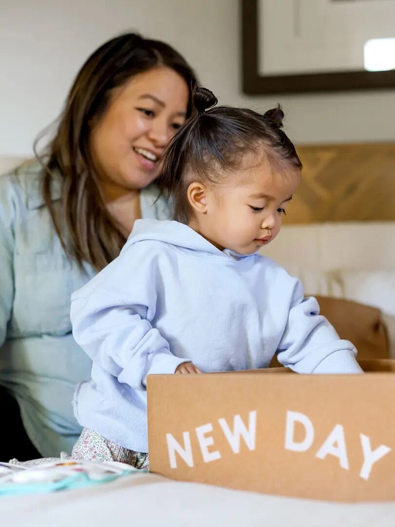 A woman sits on a bed watching a young child who is looking into an open cardboard box labeled "NEW DAY.