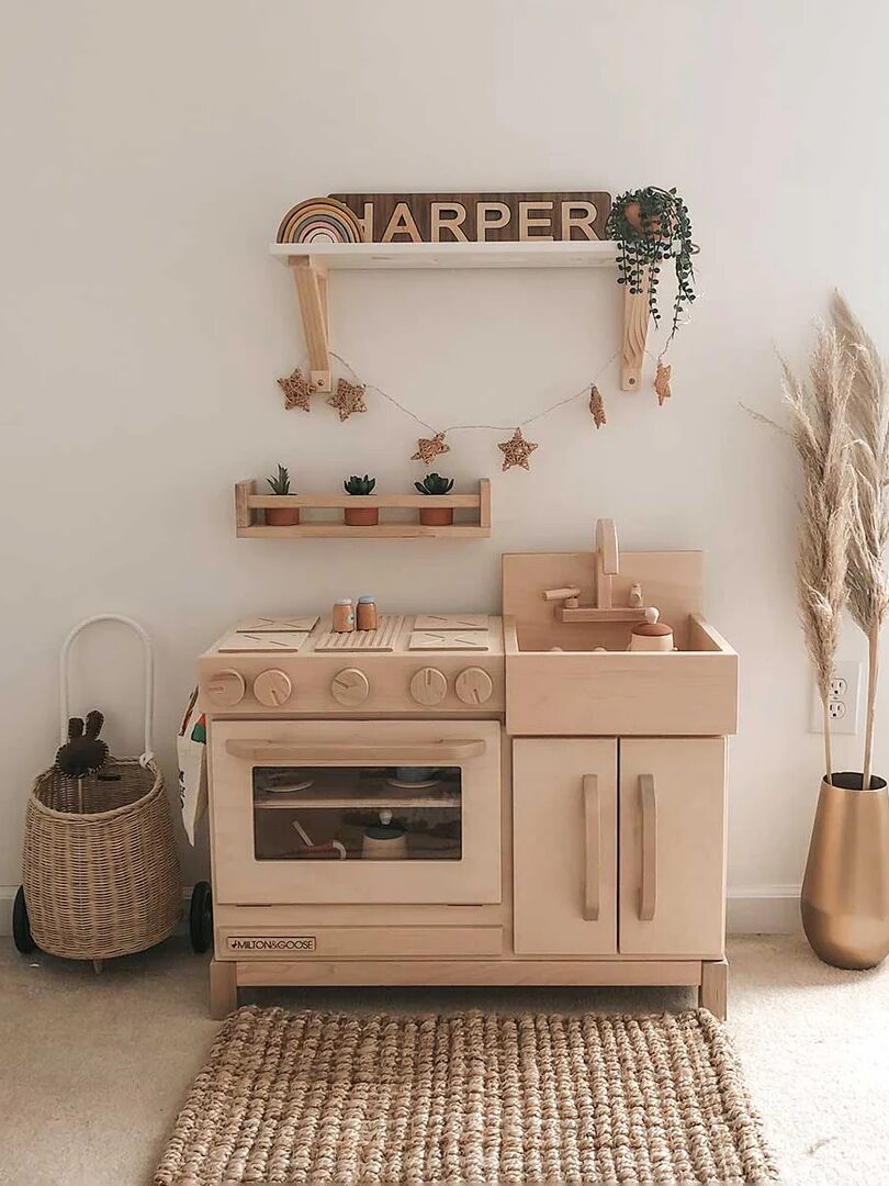 A wooden toy kitchen set is placed against a white wall, with woven baskets, a jute rug, and a gold vase with dried grass nearby. A shelf above holds decor and a name sign reading "Harper.