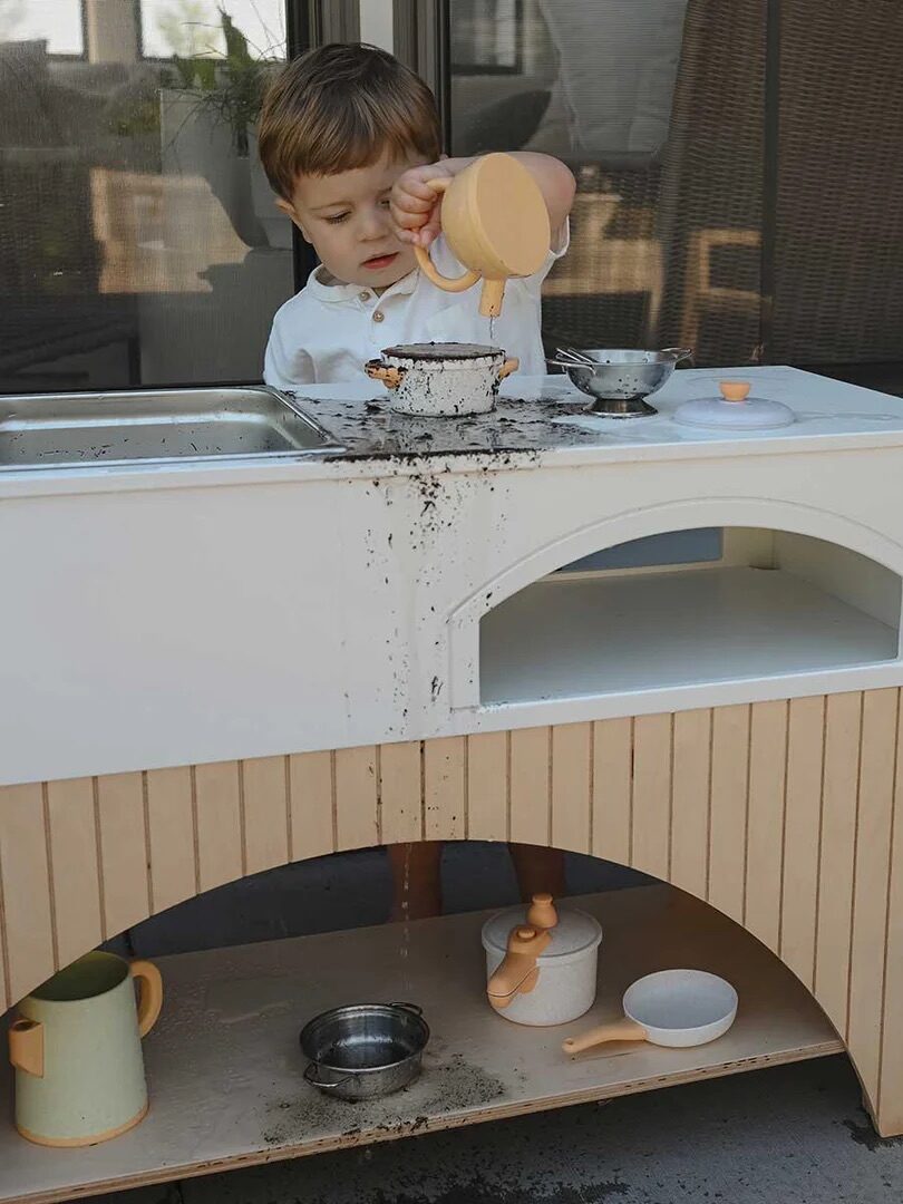 A young child pours soil from a cup onto a small table, spilling it over the edge. Various kitchen play items and a potted plant are nearby.