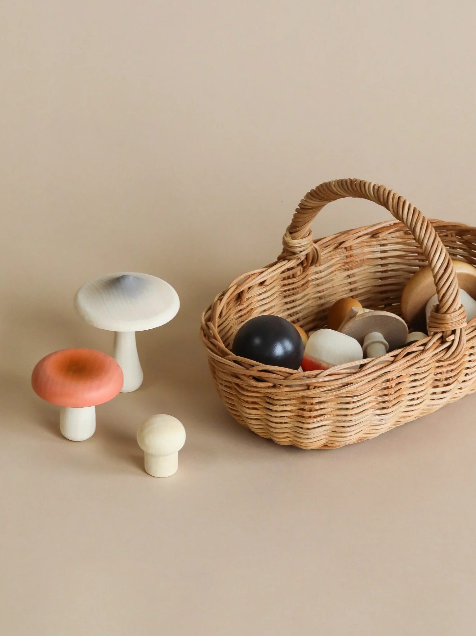 A wicker basket filled with wooden toy shapes sits on a beige surface next to three wooden mushroom figures in white, orange, and gray.