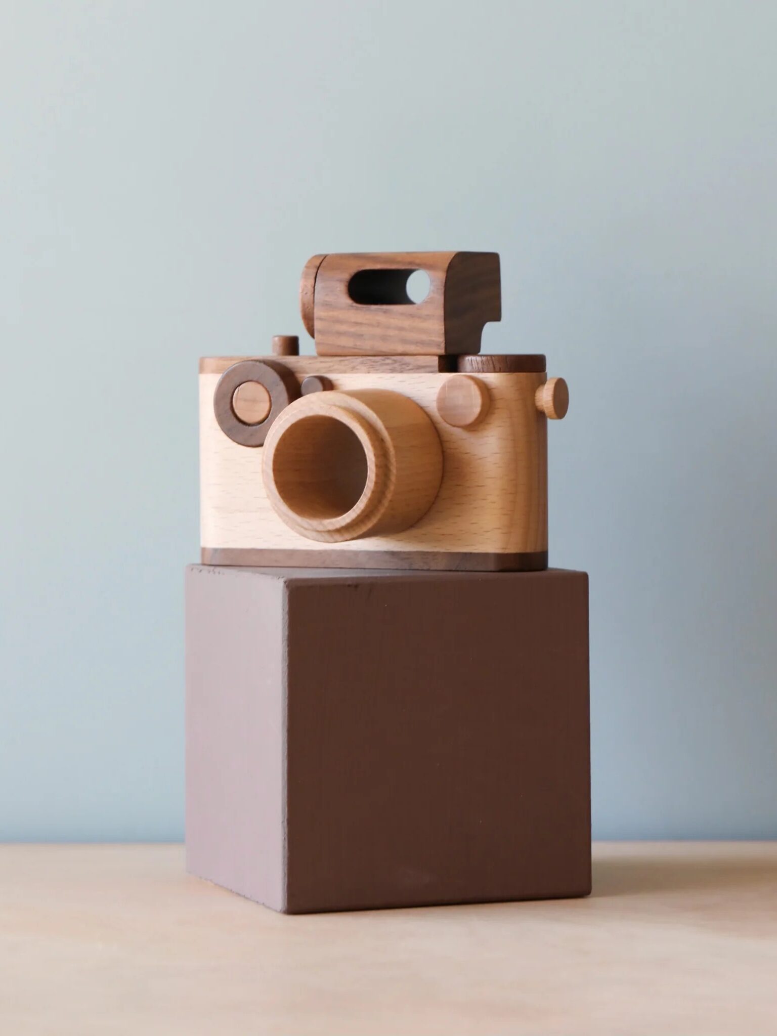 A wooden toy camera with simple geometric shapes sits on a brown rectangular block against a plain light background.