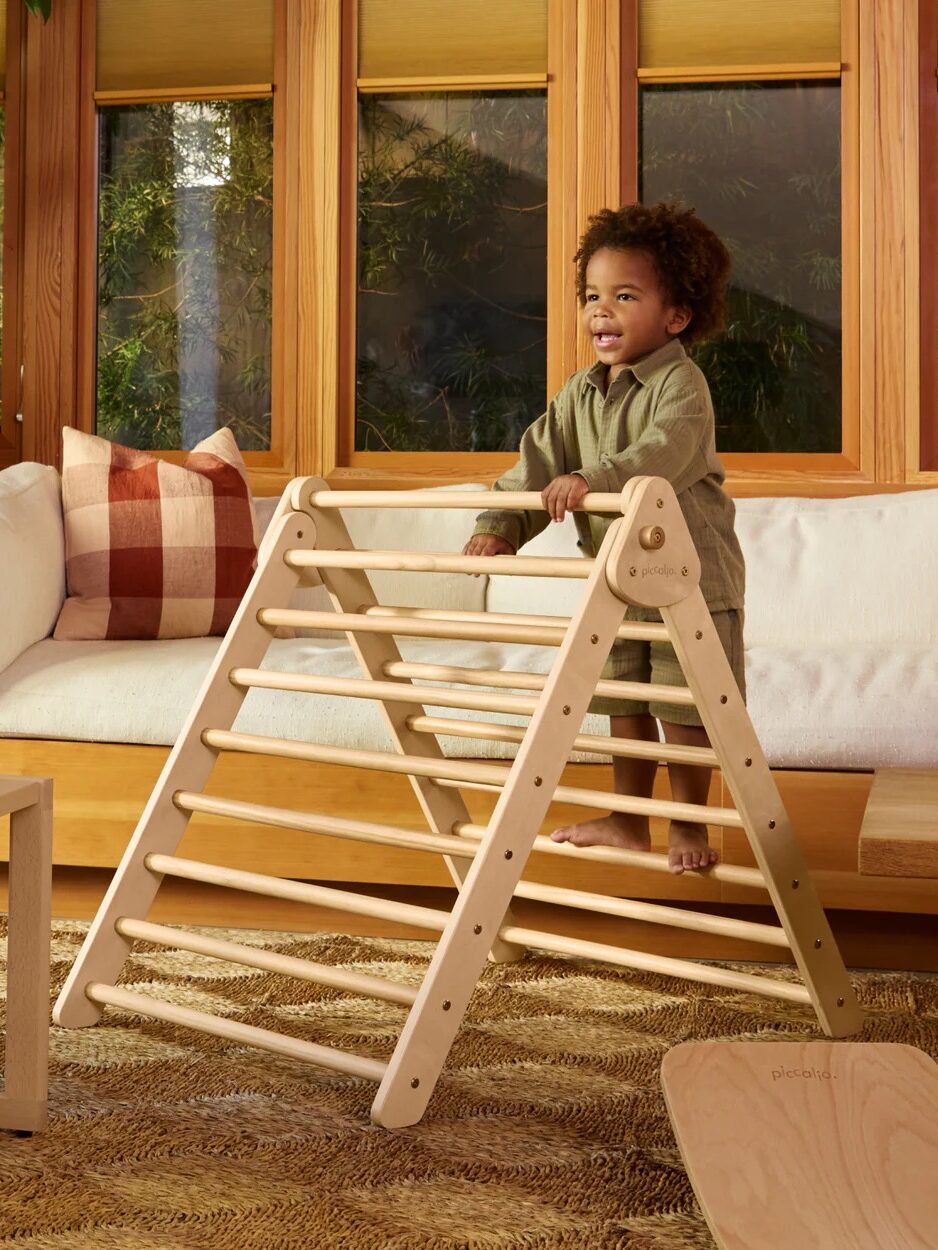 A young child stands and climbs on a wooden indoor play structure in a living room with large windows and a sofa in the background.