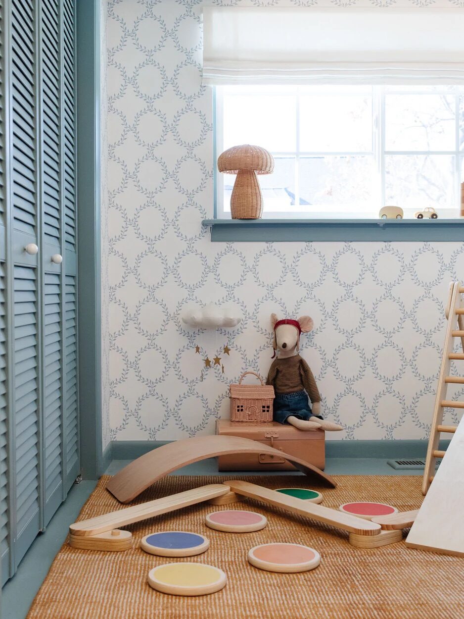 A children's playroom with patterned wallpaper, a toy mouse, a wicker lamp, wooden climbing toys, and colored stepping stones on a woven rug near a window.