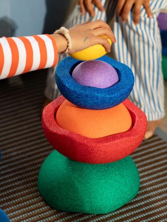 A child’s hand stacks colorful, textured foam shapes on a mat, with another child sitting nearby in striped pants.