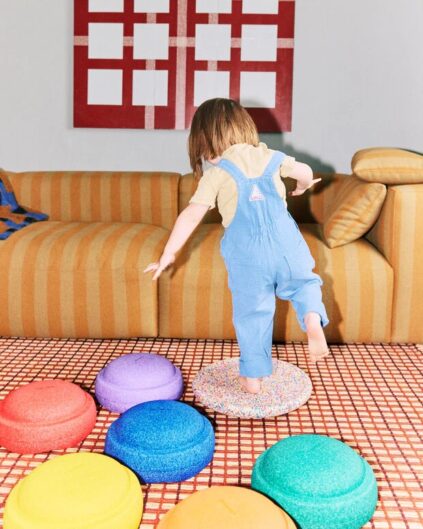 A young child in blue overalls balances on a textured mat, surrounded by colorful padded cushions in a living room with a striped sofa and geometric wall art.