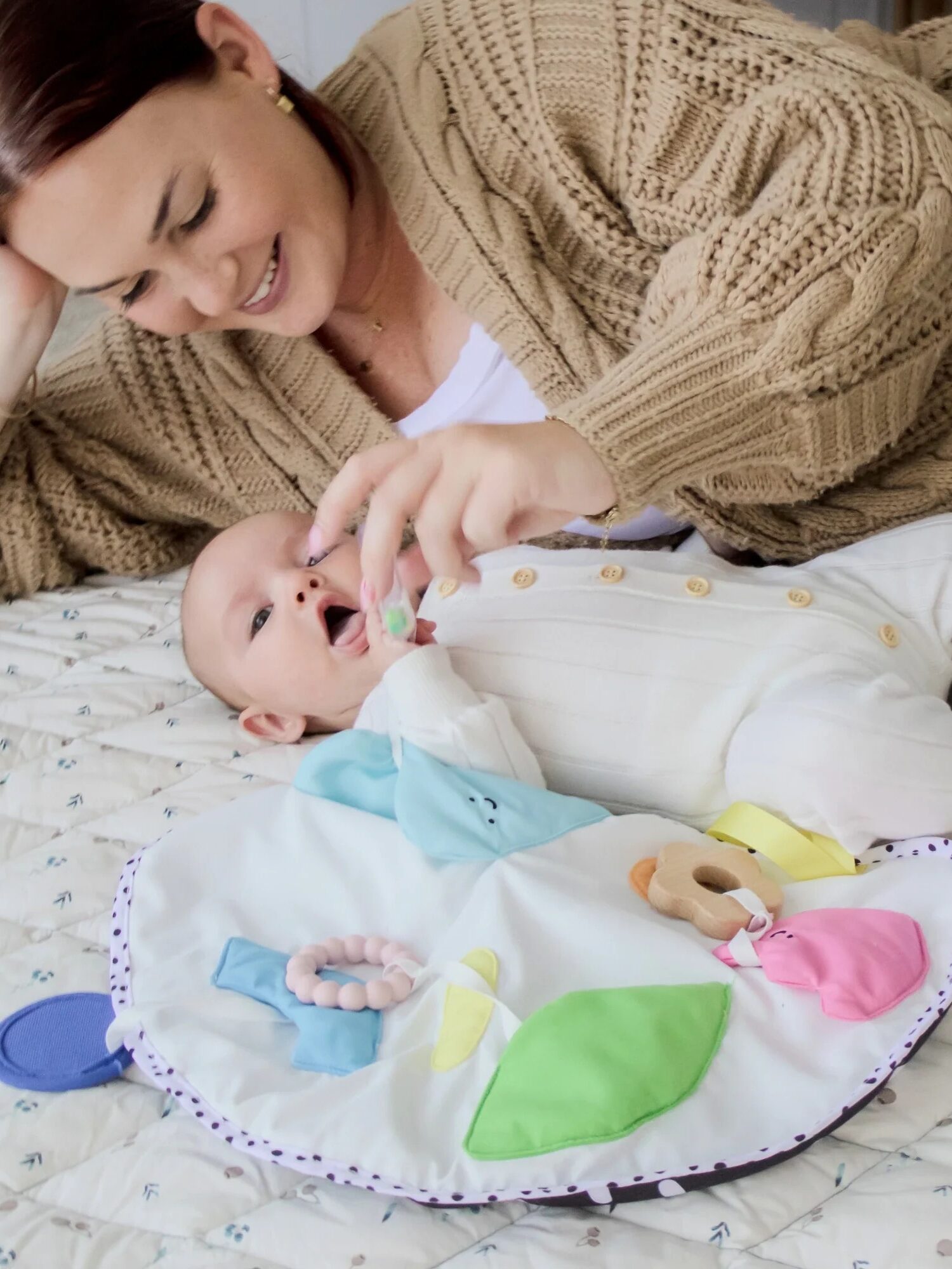 An adult lies on a bed, smiling and engaging with a baby who is lying on a quilt, reaching for soft toys on a colorful play mat.