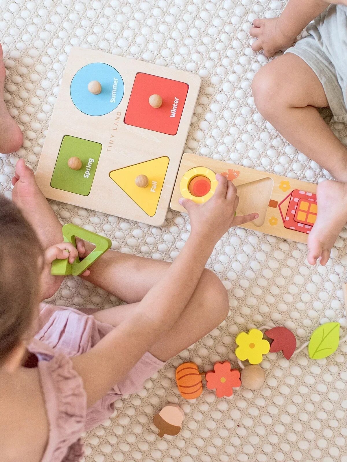 Two young children sit on a textured rug, playing with colorful wooden puzzles and shape toys, sorting pieces by season and shape.