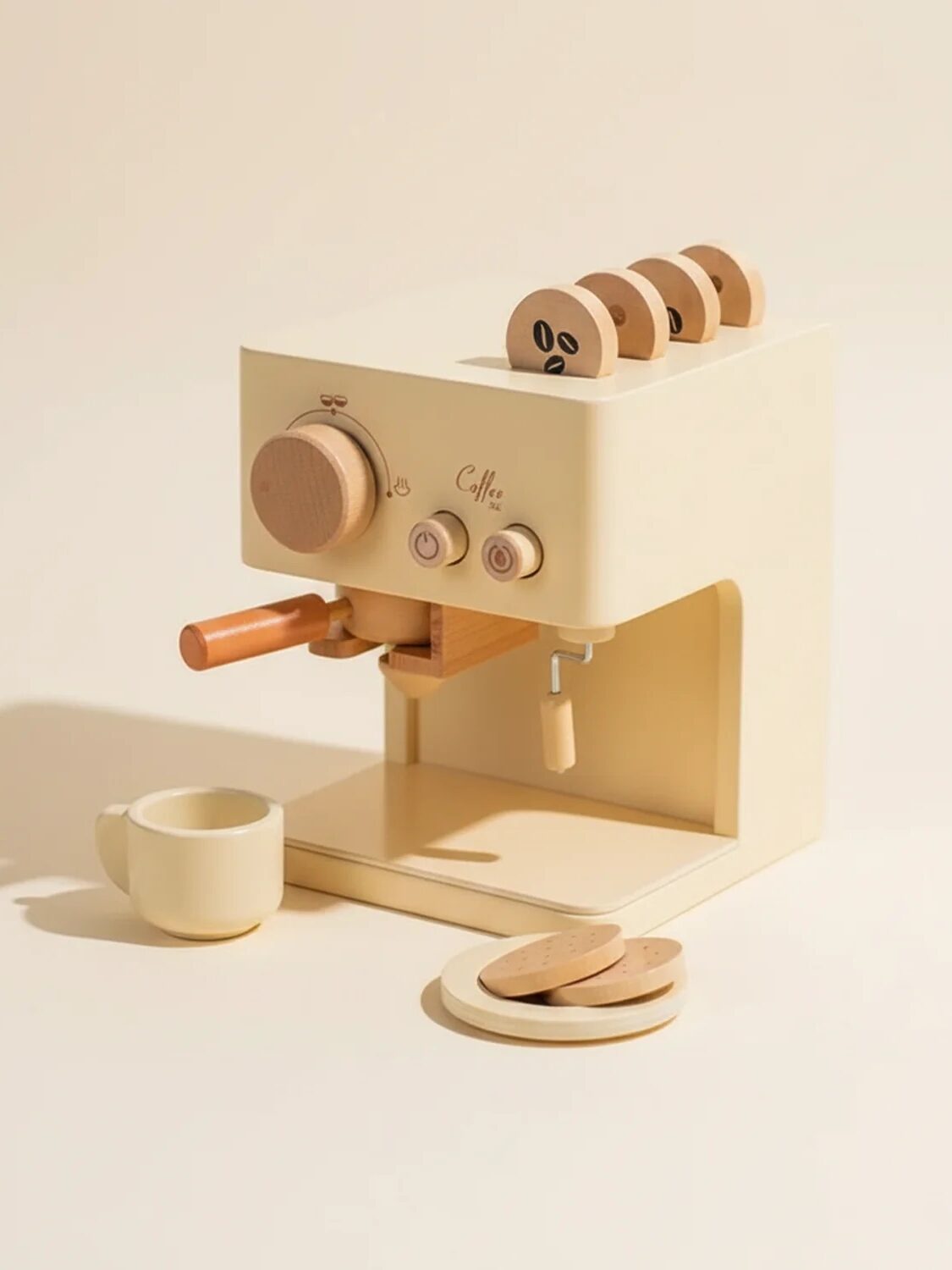A light beige toy coffee machine with wooden knobs and accessories, including a cup and saucers, is displayed against a neutral background.