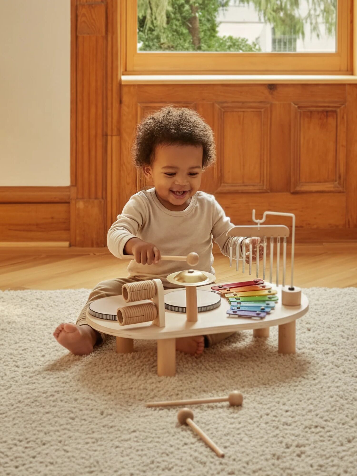 A young child sits on a carpeted floor playing with a wooden musical toy set in a sunlit room with wooden paneled walls and a large window.