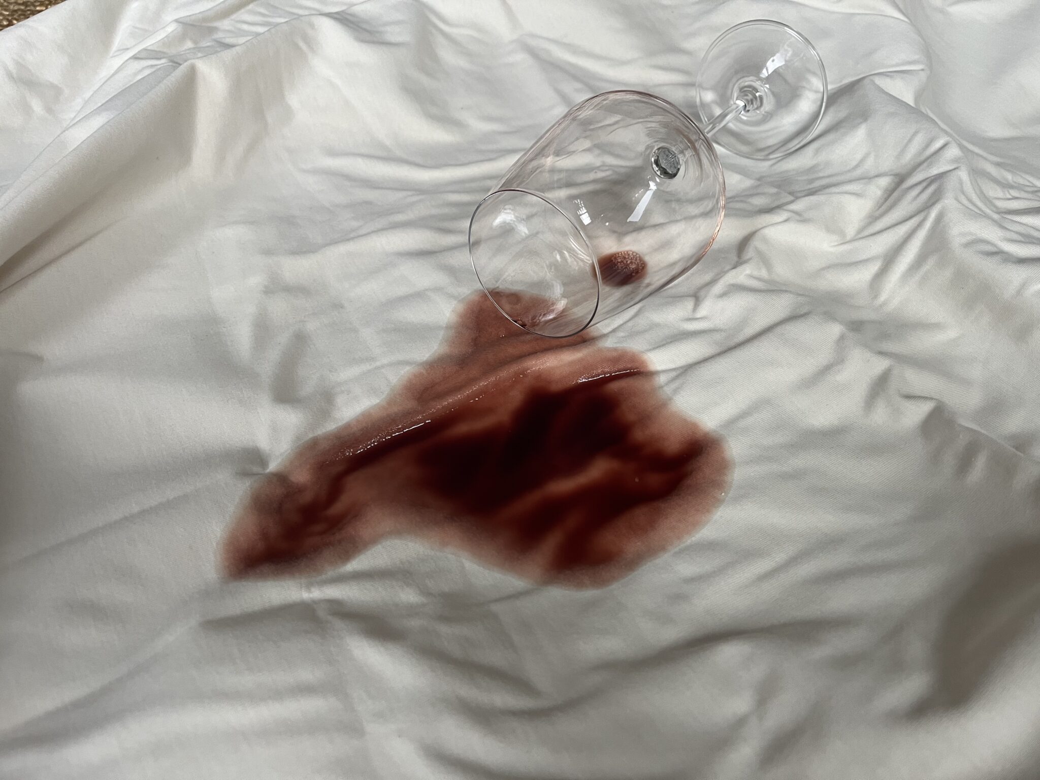 A wine glass tipped over on a white sheet, spilling red wine and creating a large stain on the fabric.
