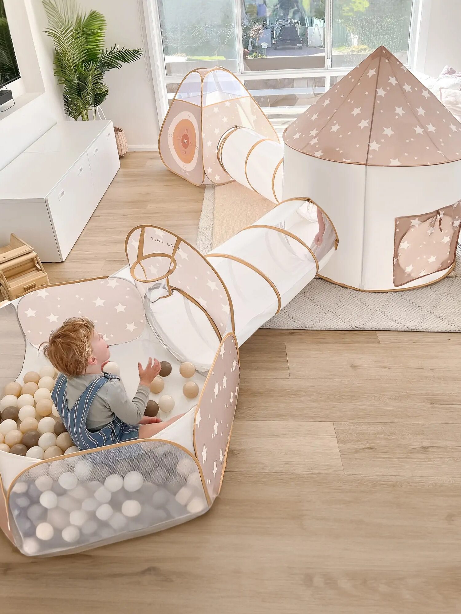 A toddler plays in a beige and white star-patterned indoor play tent set with a tunnel and ball pit in a bright living room.