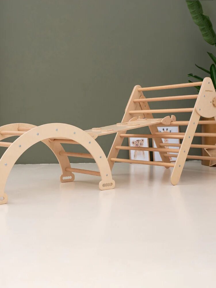 A wooden indoor climbing structure for children, featuring an arch, a ramp, and a triangle frame, sits on a light floor with a green wall and plant in the background.