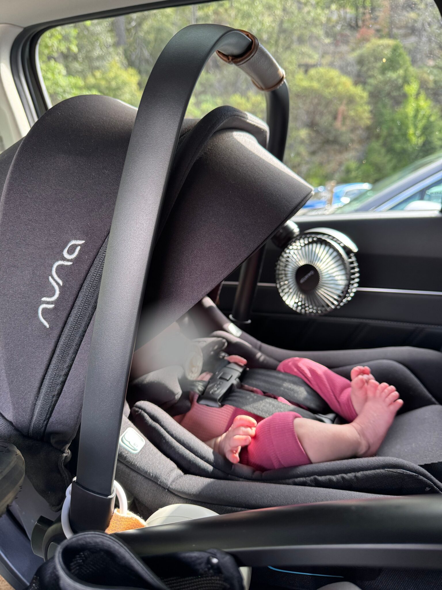 A baby in a Nuna car seat with a small fan attached to the headrest, positioned in the backseat of a car.