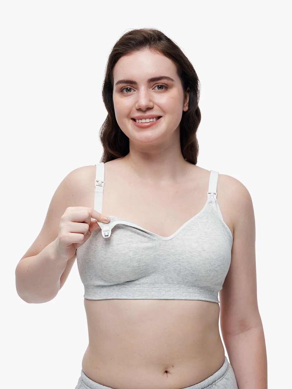 A woman wearing a light gray nursing bra smiles at the camera while unclipping one cup to demonstrate its breastfeeding feature.