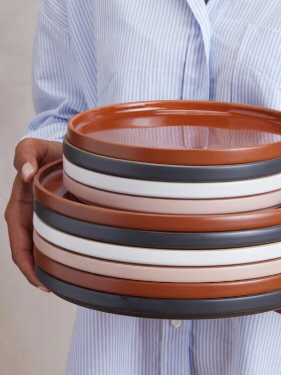 A person wearing a blue and white striped shirt holds a stack of round ceramic plates in brown, white, and dark gray colors.