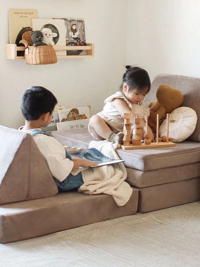 Two young children sit in a cozy playroom with soft seating, wooden toys, and a table. One child reads a book while the other plays with blocks. The space is decorated in neutral tones.