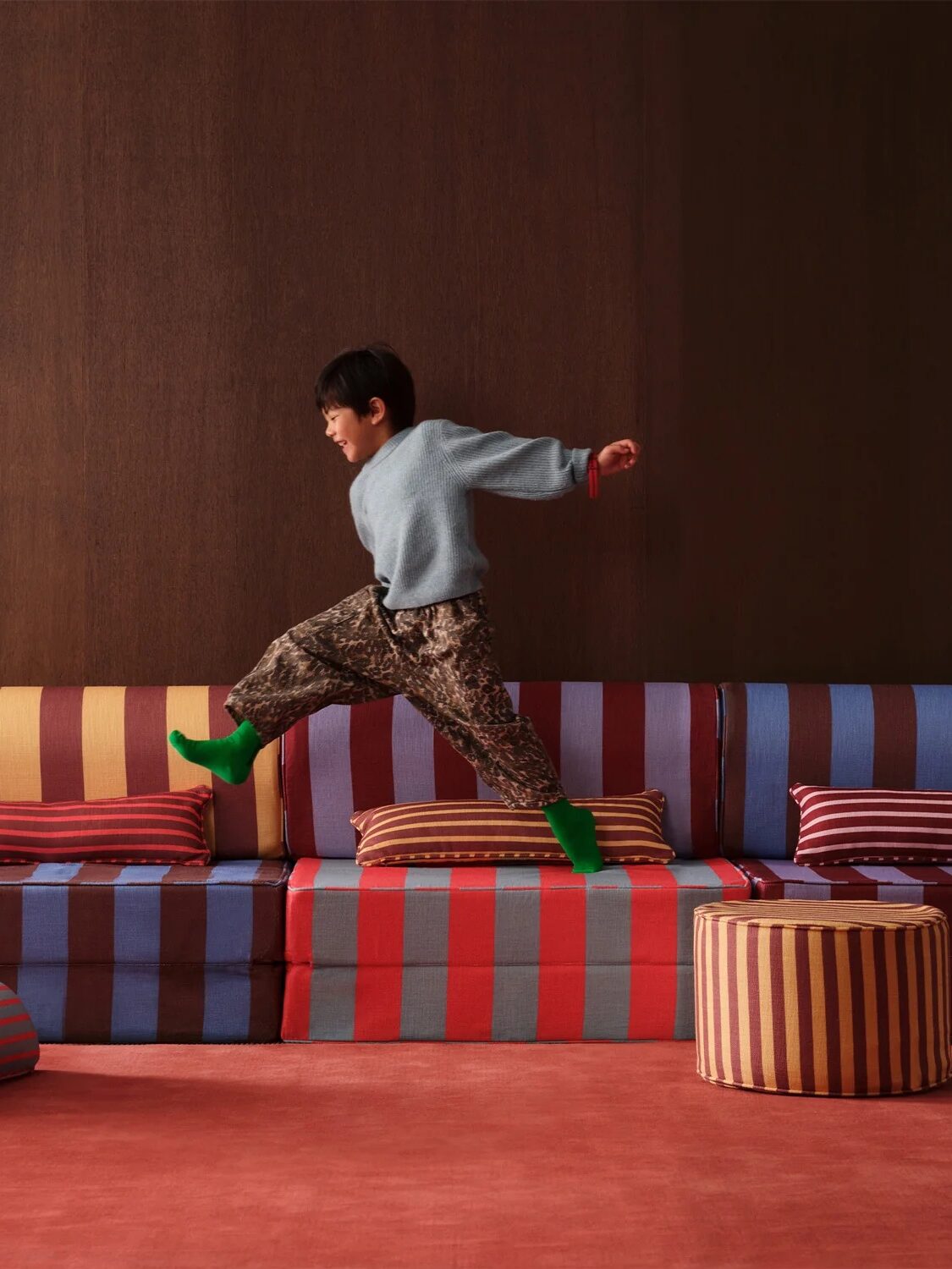 A child in a gray sweater and patterned pants jumps across a striped, colorful modular sofa set in a room with a dark wall and red carpet.