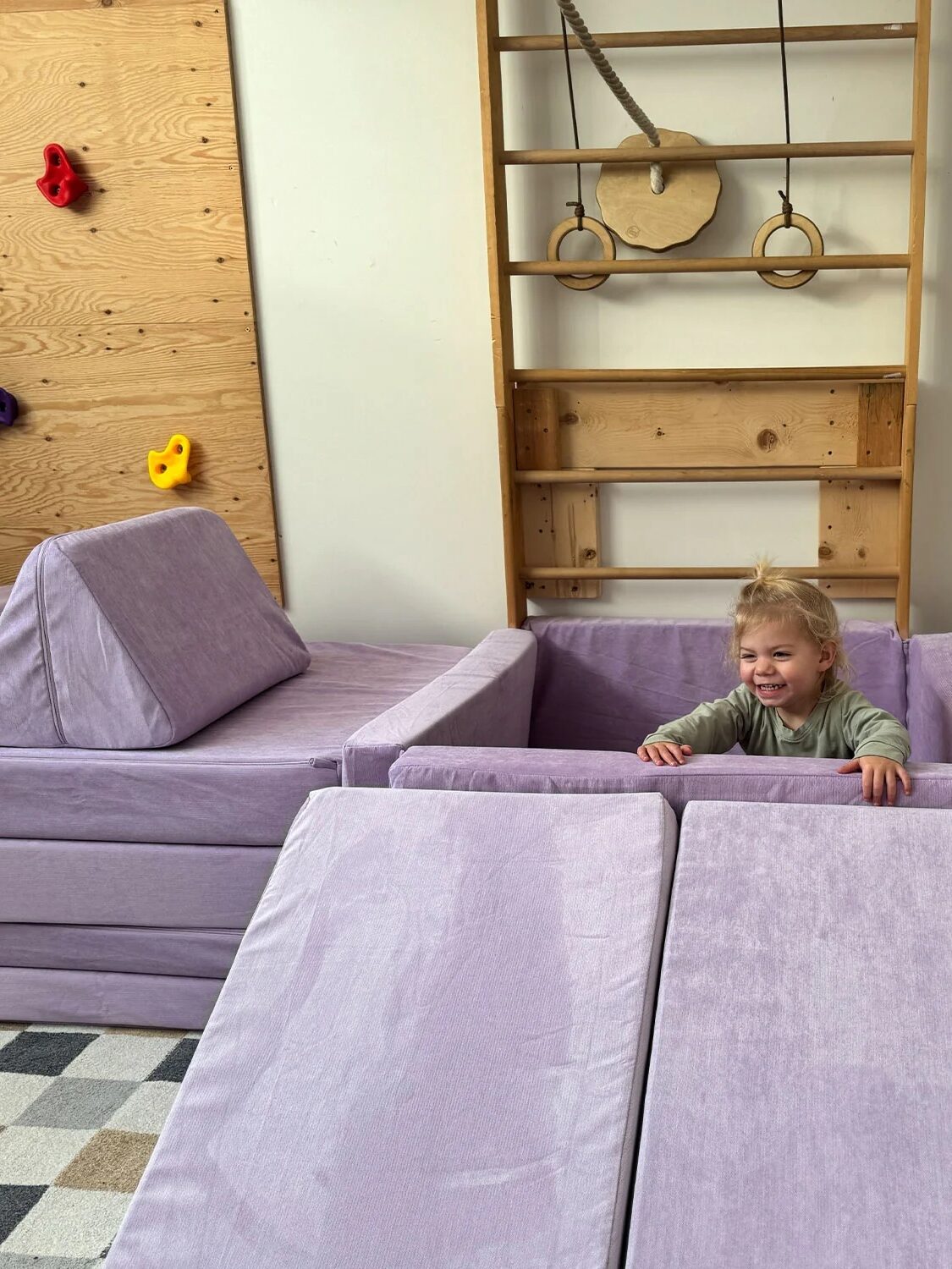 A young child smiles while playing in a soft, light purple foam playset in a playroom with a wooden climbing wall and gym equipment.