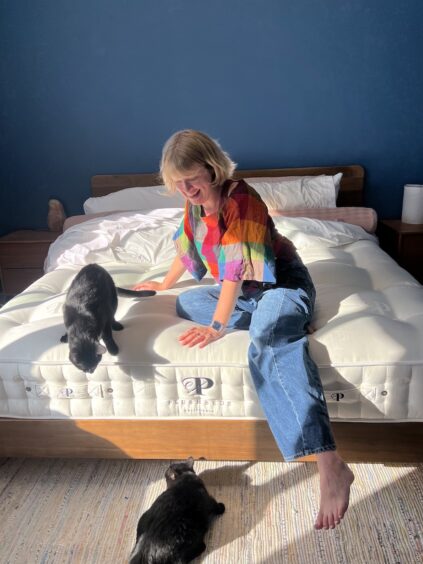 A woman sits on a bed in a sunlit room, smiling at two black cats; one cat is on the bed and the other is on the floor.