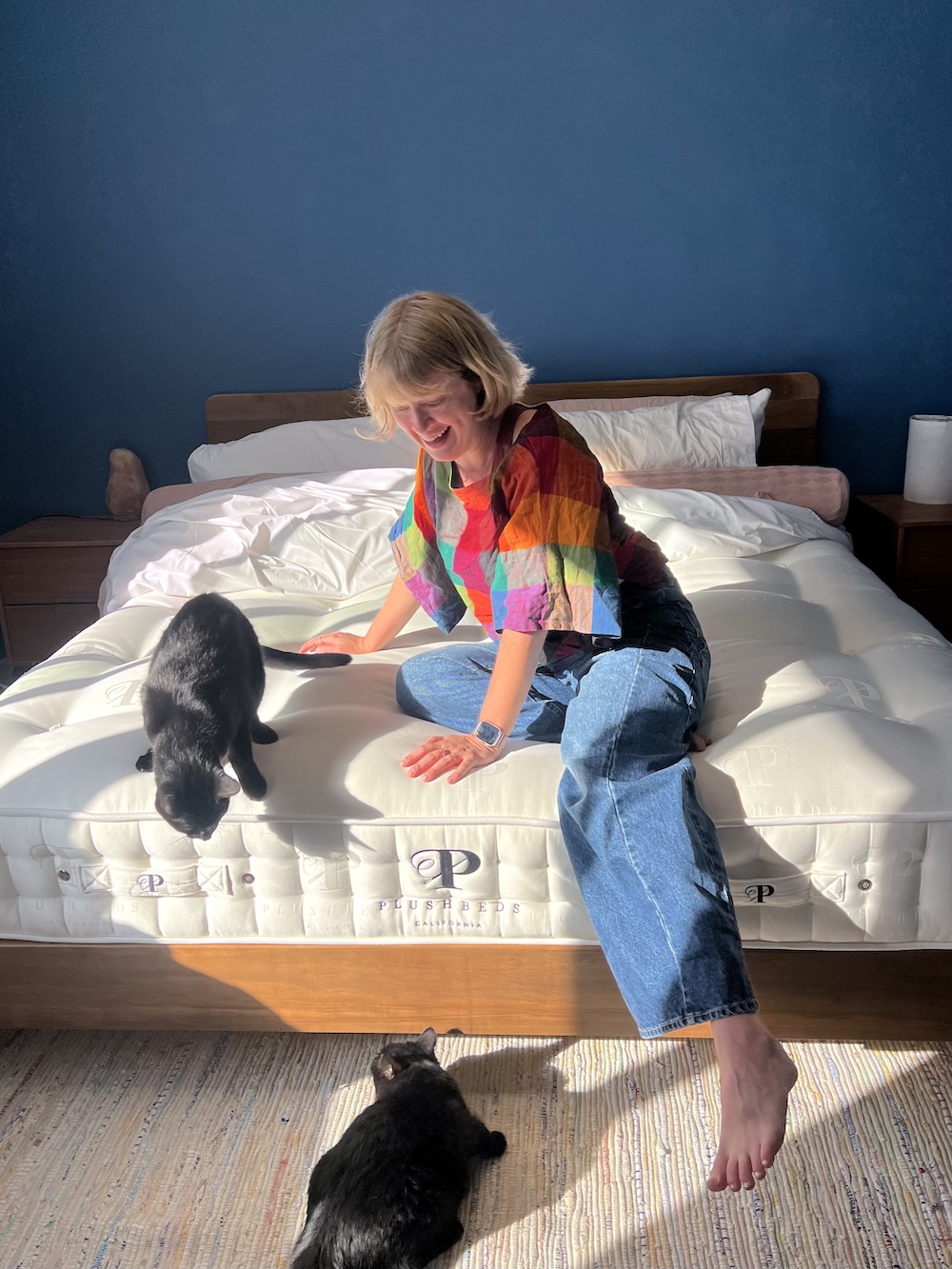 A woman sits on a bed in a sunlit room, smiling at two black cats; one cat is on the bed and the other is on the floor.