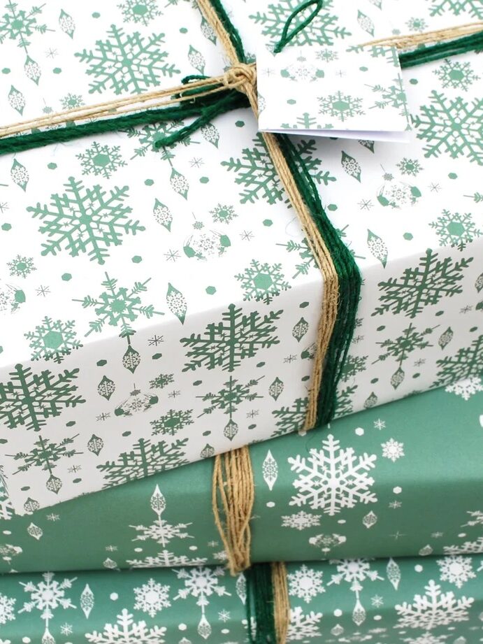 Three gift boxes wrapped in green and white snowflake-patterned paper, stacked and tied with brown string, with a blank tag attached.