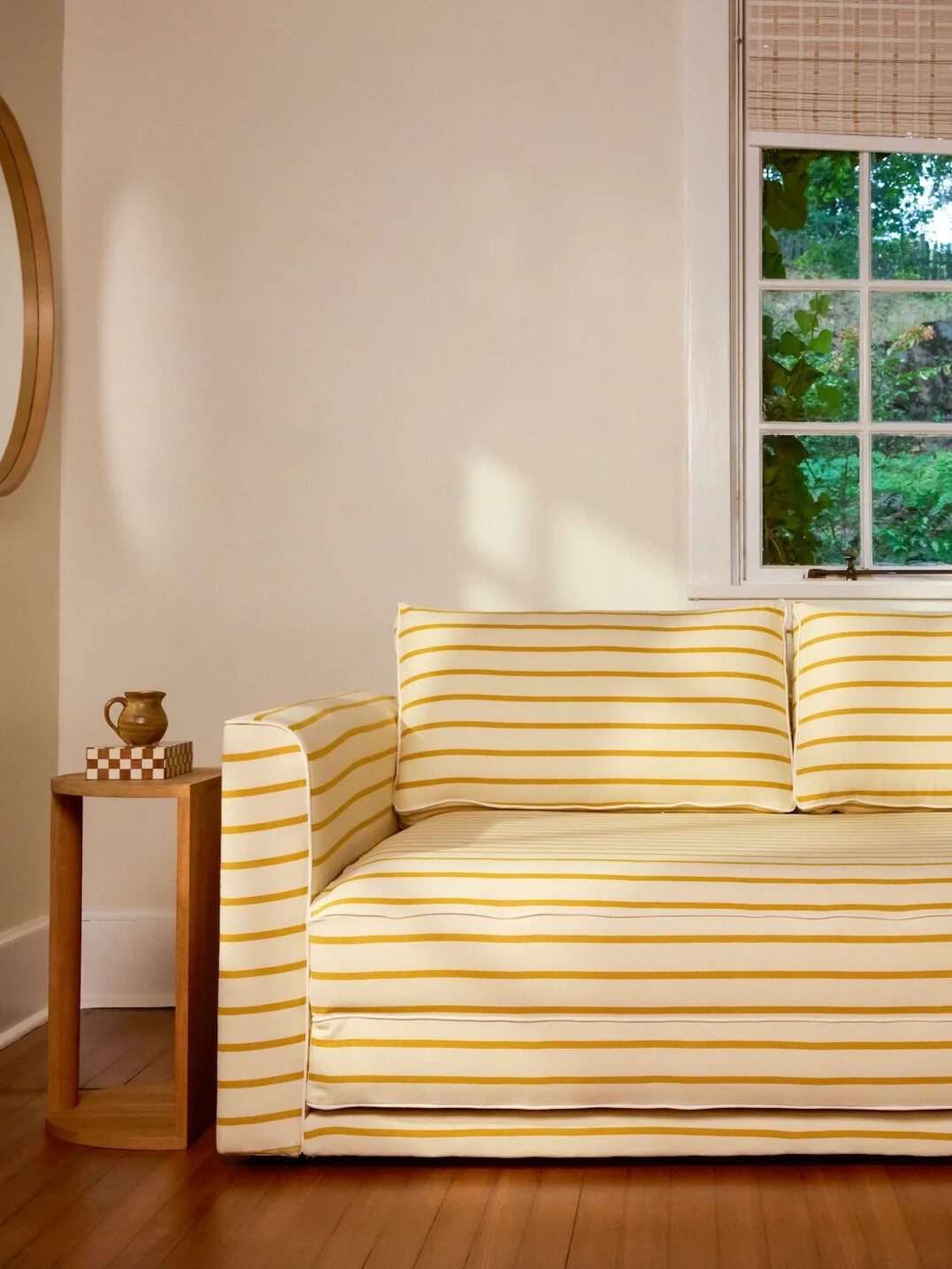 A yellow and white striped sofa with two cushions is placed against a wall, with a small round side table and a beige throw blanket, in front of windows with woven blinds.