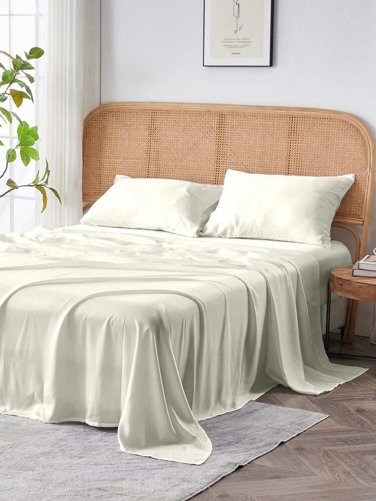 A made bed with white sheets and two pillows, rattan headboard, potted plant by a window, and a small round wooden side table on a gray rug.