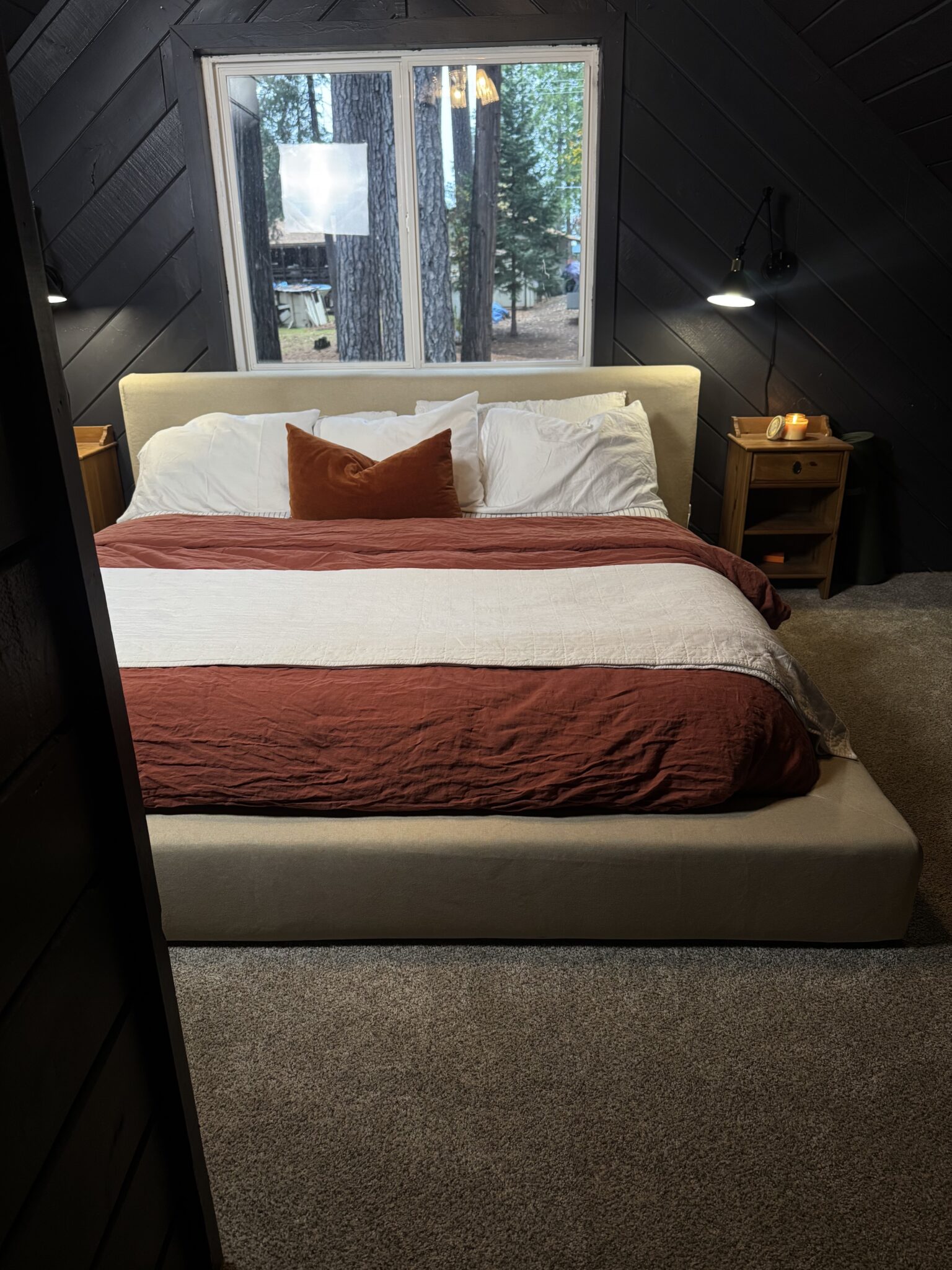 A neatly made bed with white and rust-colored bedding, two nightstands with lamps, set in a carpeted room with a large window overlooking trees.