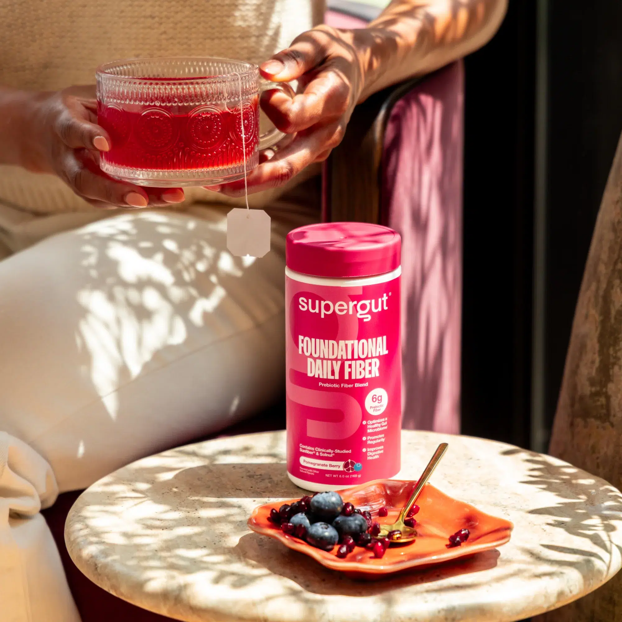 A person holds a glass cup with red tea and a tea bag, next to a container of Supergut daily fiber powder—one of the top fiber supplements for women—alongside a plate of berries on an orange dish.