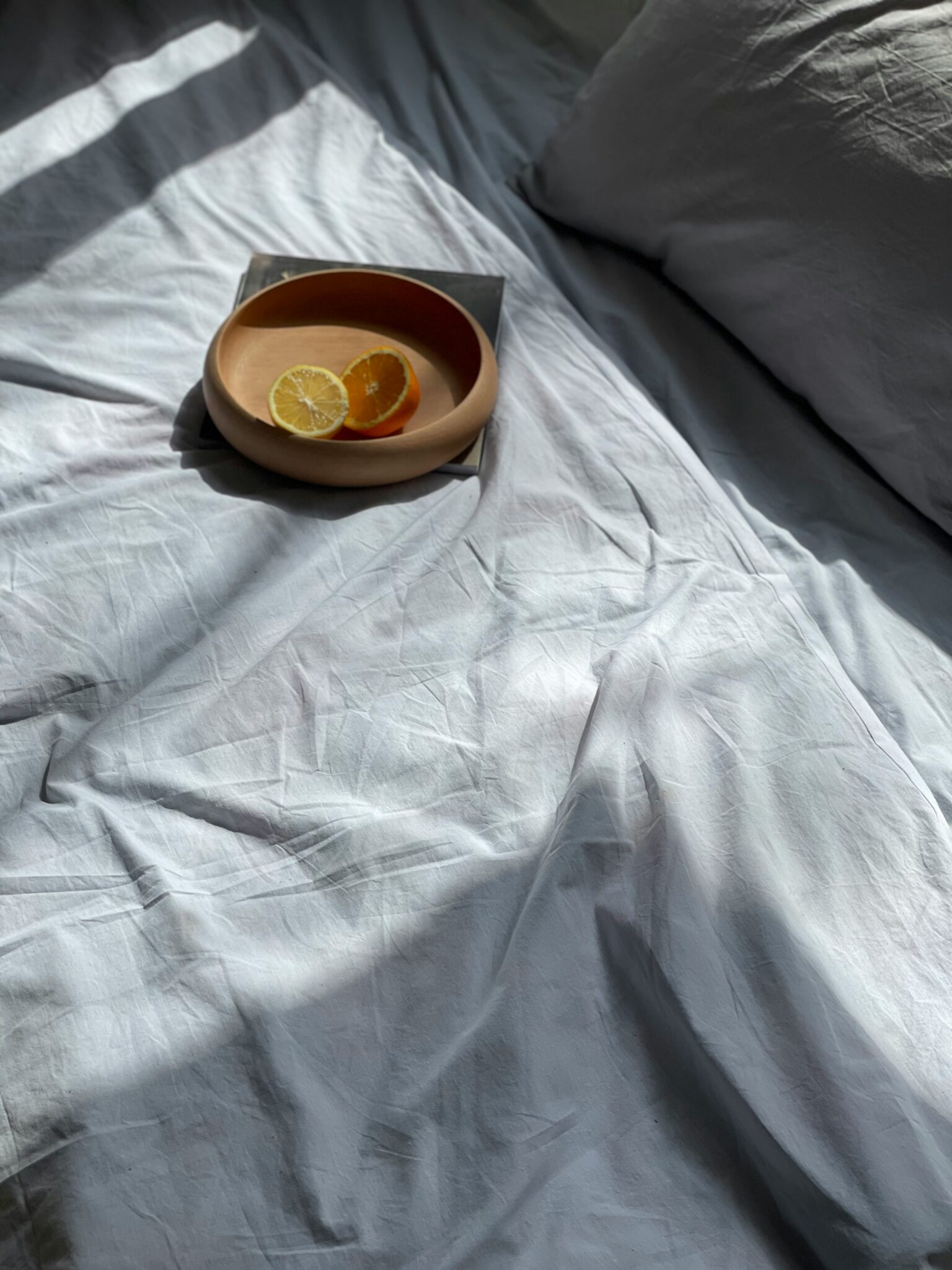 A round wooden plate with lemon and orange slices rests on a wrinkled white bedsheet next to a pillow, under natural light.