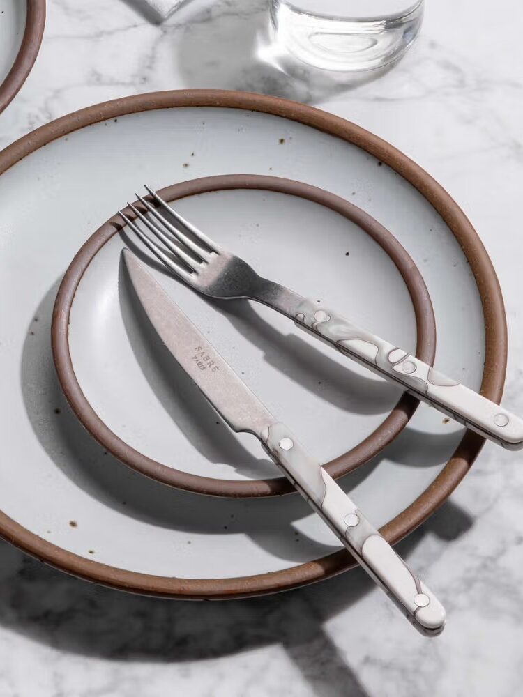 A set of ceramic plates with brown rims stacked on a marble surface, with a fork and knife placed diagonally on top.