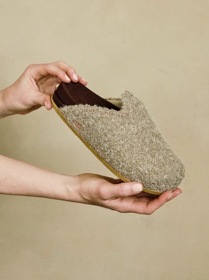 A person holds a pair of grey woolen slippers with a brown interior and a light brown sole against a plain beige background.